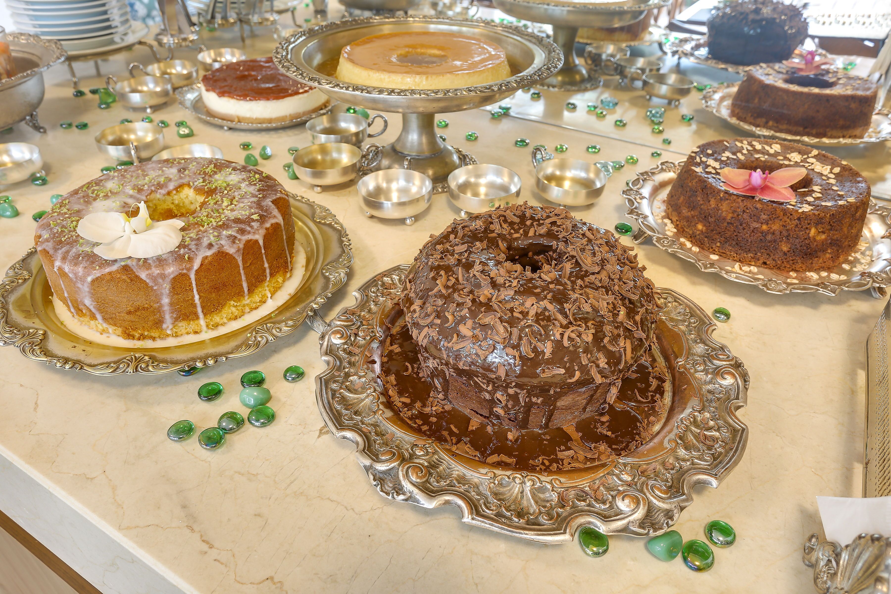 Una mesa con varios pasteles sobre pedestales de plata ornamentados, rodeada de pequeños cuencos y elementos decorativos verdes.