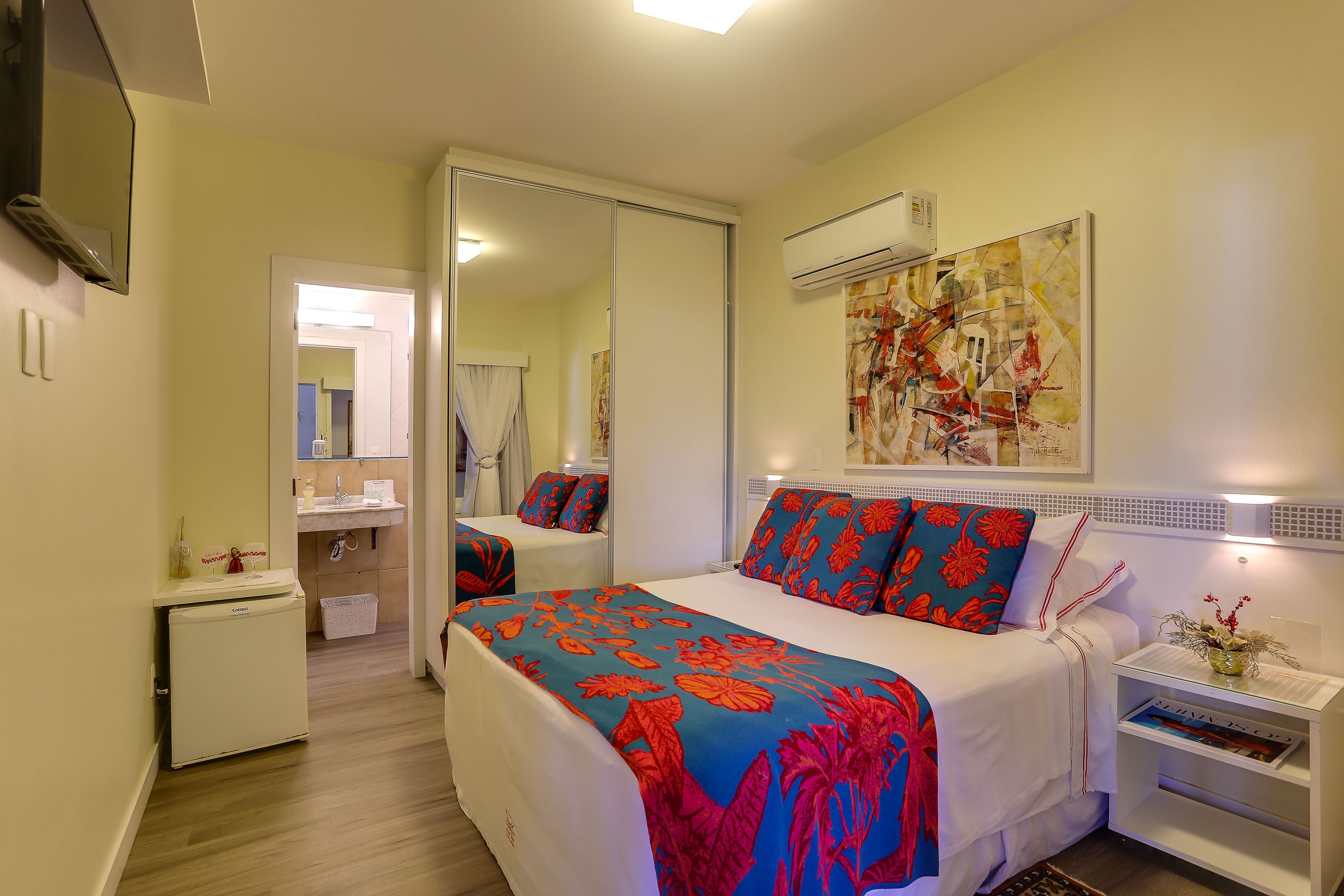 Habitación de hotel con cama, mesita de noche pequeña, espejo, aire acondicionado montado en la pared y puerta que da a un baño.