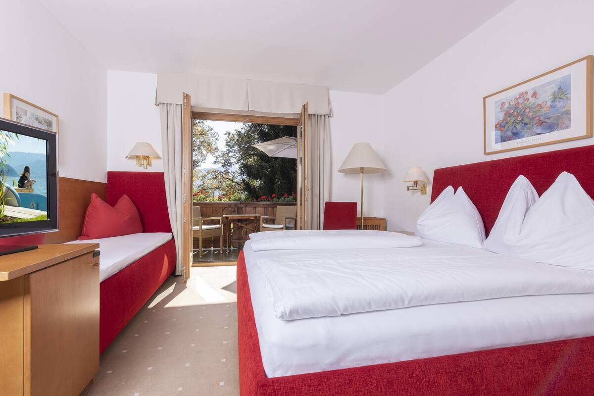 Ein Hotelzimmer mit Bett, Schreibtisch, Fernseher, einem Balkon mit Tisch und Stühlen sowie einem Bild an der Wand.