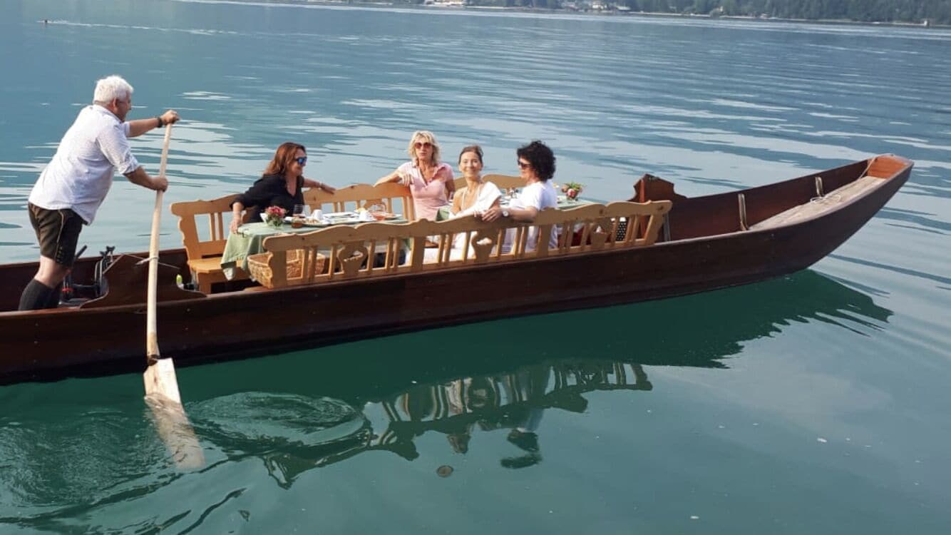 A wooden boat with a table and chairs, people seated, and a person rowing.