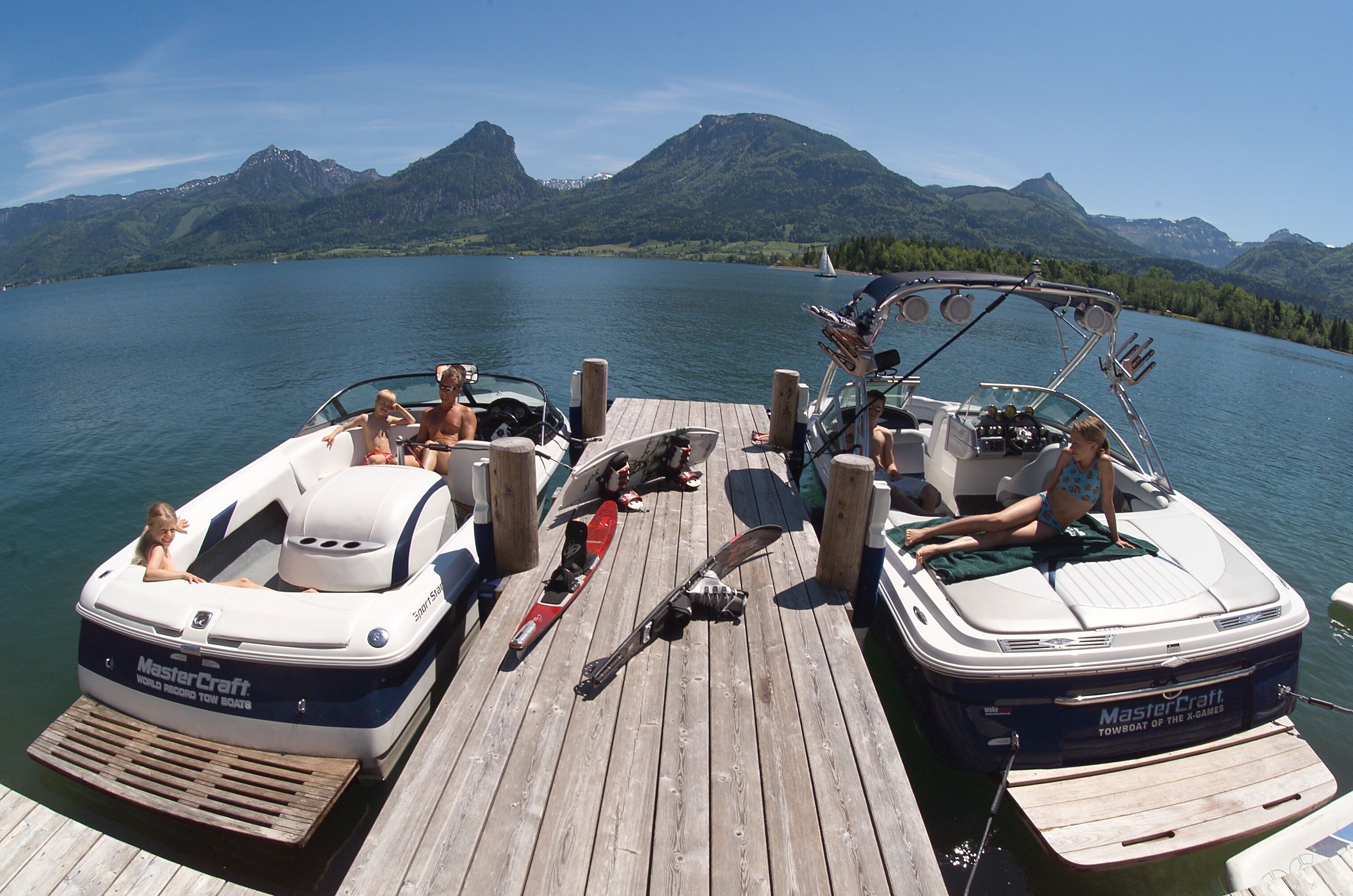 A wooden dock with two boats, one with skis and another with a canopy, docked at a lake with mountains in the background.