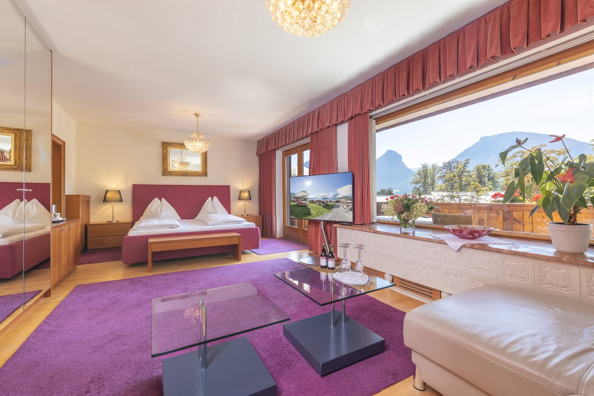 Ein modernes Hotelzimmer mit Bett, Sofa, Glastisch und einem großen Fenster mit Blick auf die Berge.