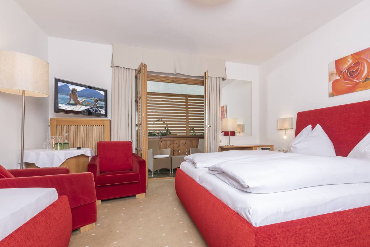 Ein Hotelzimmer mit einem Bett, einem roten Sessel, einem Schreibtisch mit Lampe, einem Fernseher und einem Gemälde.