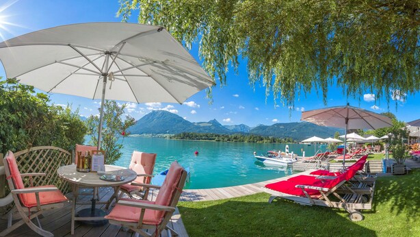A lakeside outdoor seating area with lounge chairs and umbrellas, a wooden deck, and a view of the lake and mountains. - Hotel Furian (Sankt Wolfgang im Salzkammergut)