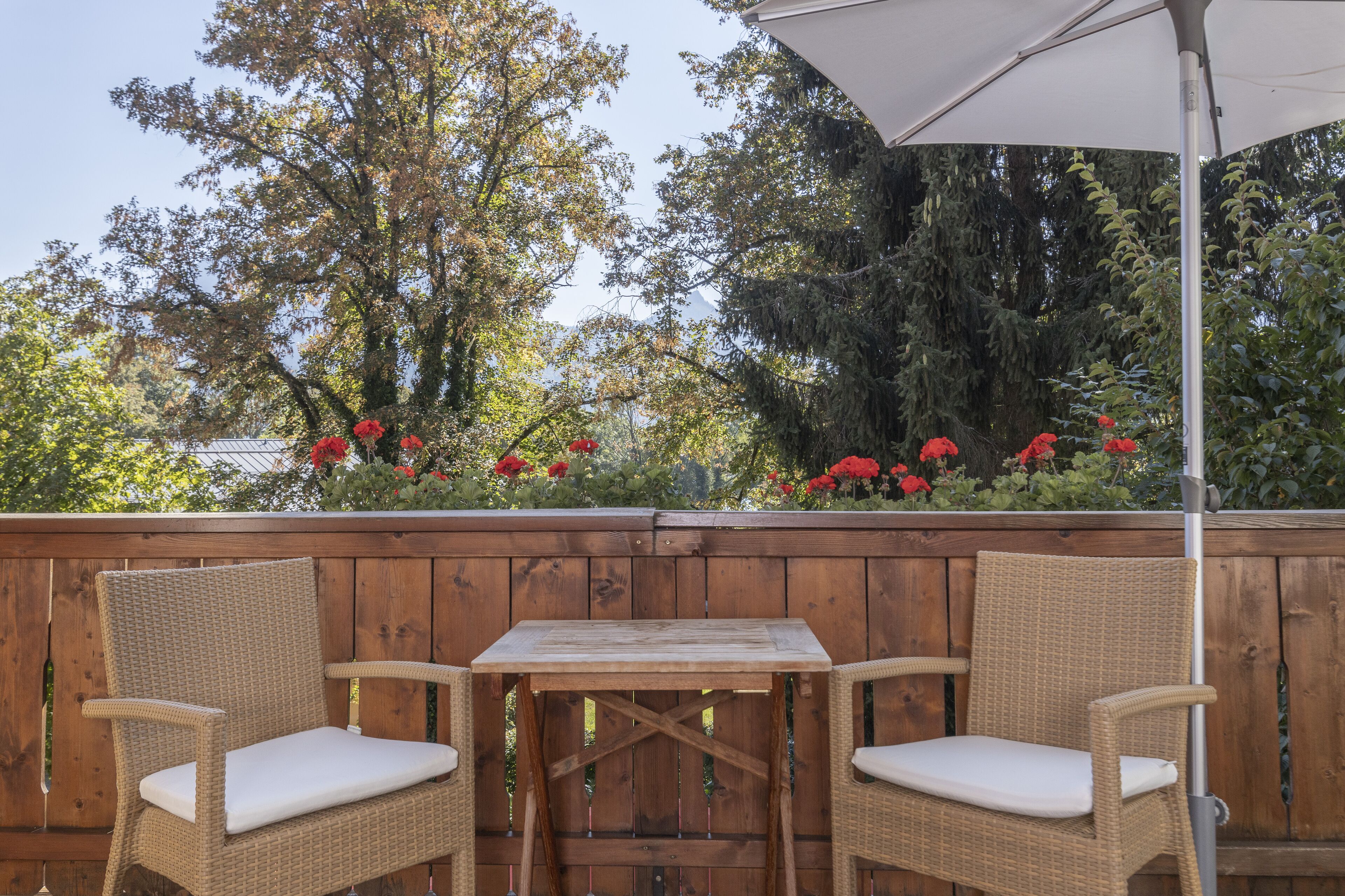 A wooden deck with wicker chairs and a table, surrounded by a wooden fence and red flowers.