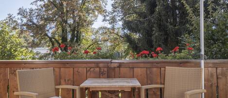 A wooden deck with wicker chairs and a table, surrounded by a wooden fence and red flowers.