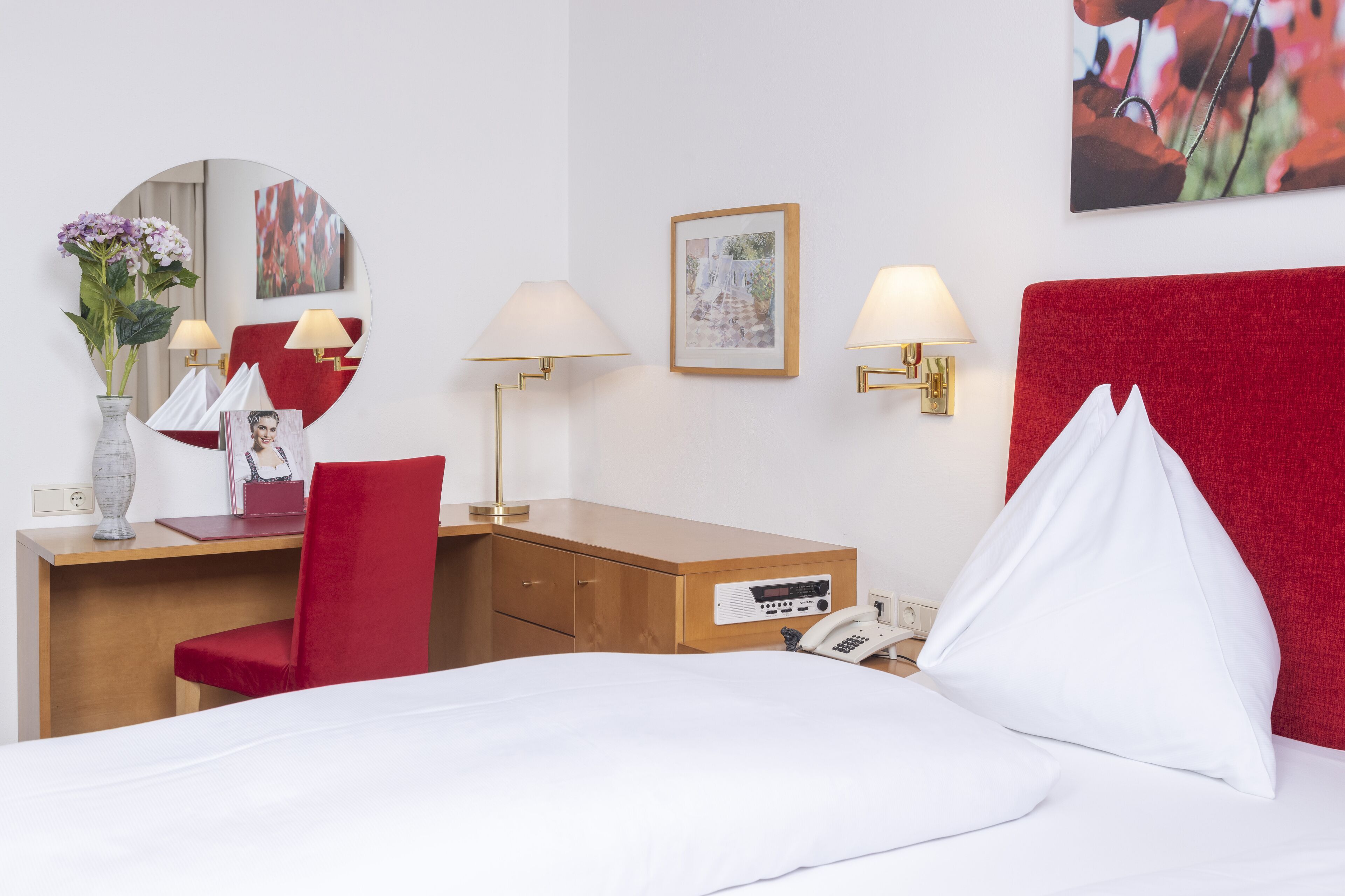 Quarto de hotel com uma cama, uma mesa com cadeira, telefone, luminária, espelho e um quadro na parede.