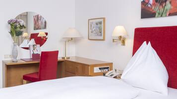 Quarto de hotel com uma cama, uma mesa com cadeira, telefone, luminária, espelho e um quadro na parede.