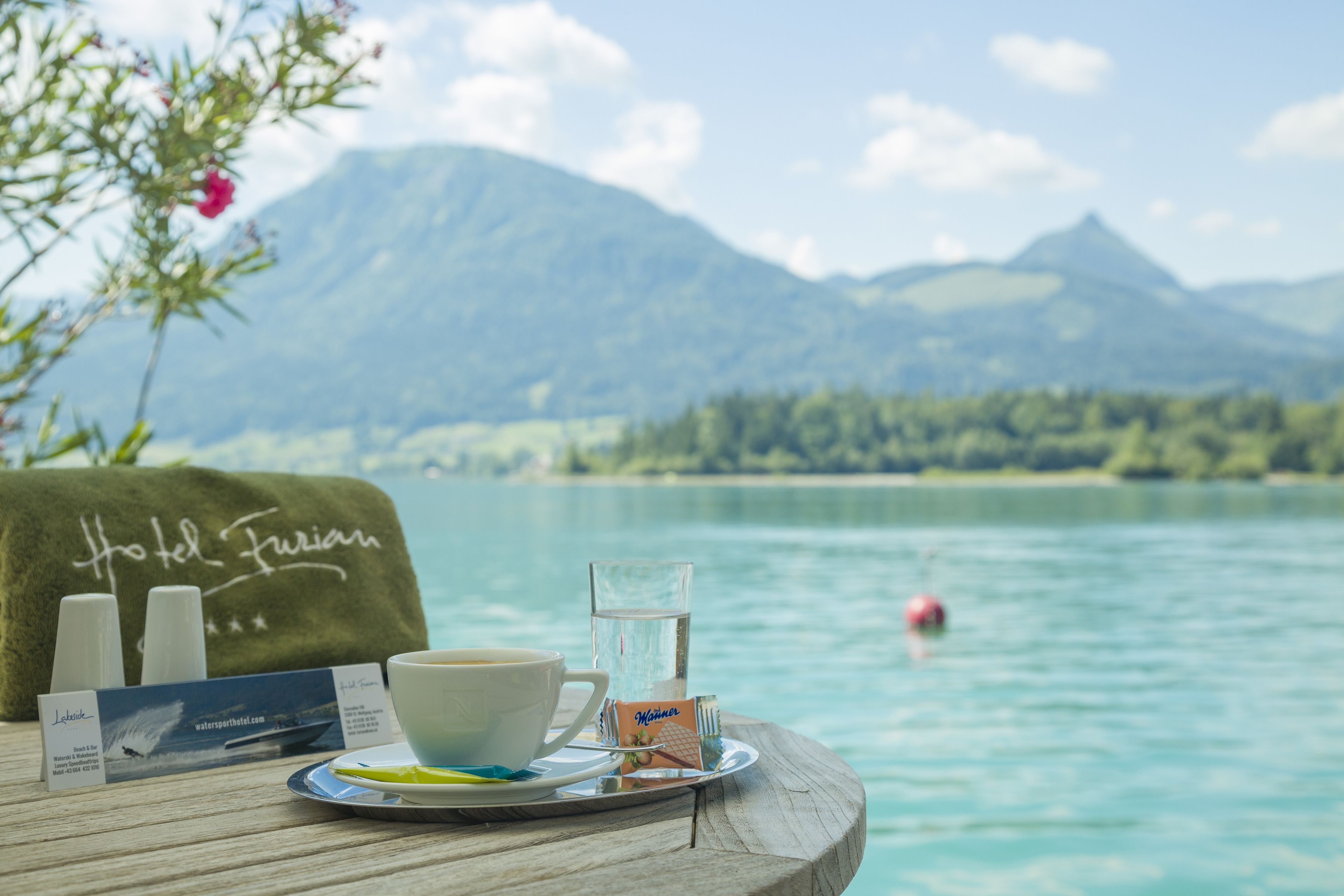 Ein Tisch mit einer Tasse Kaffee, einem Glas Wasser und einem Handtuch mit der Aufschrift "Hotel Furian", mit Blick auf einen See und Berge im Hintergrund.