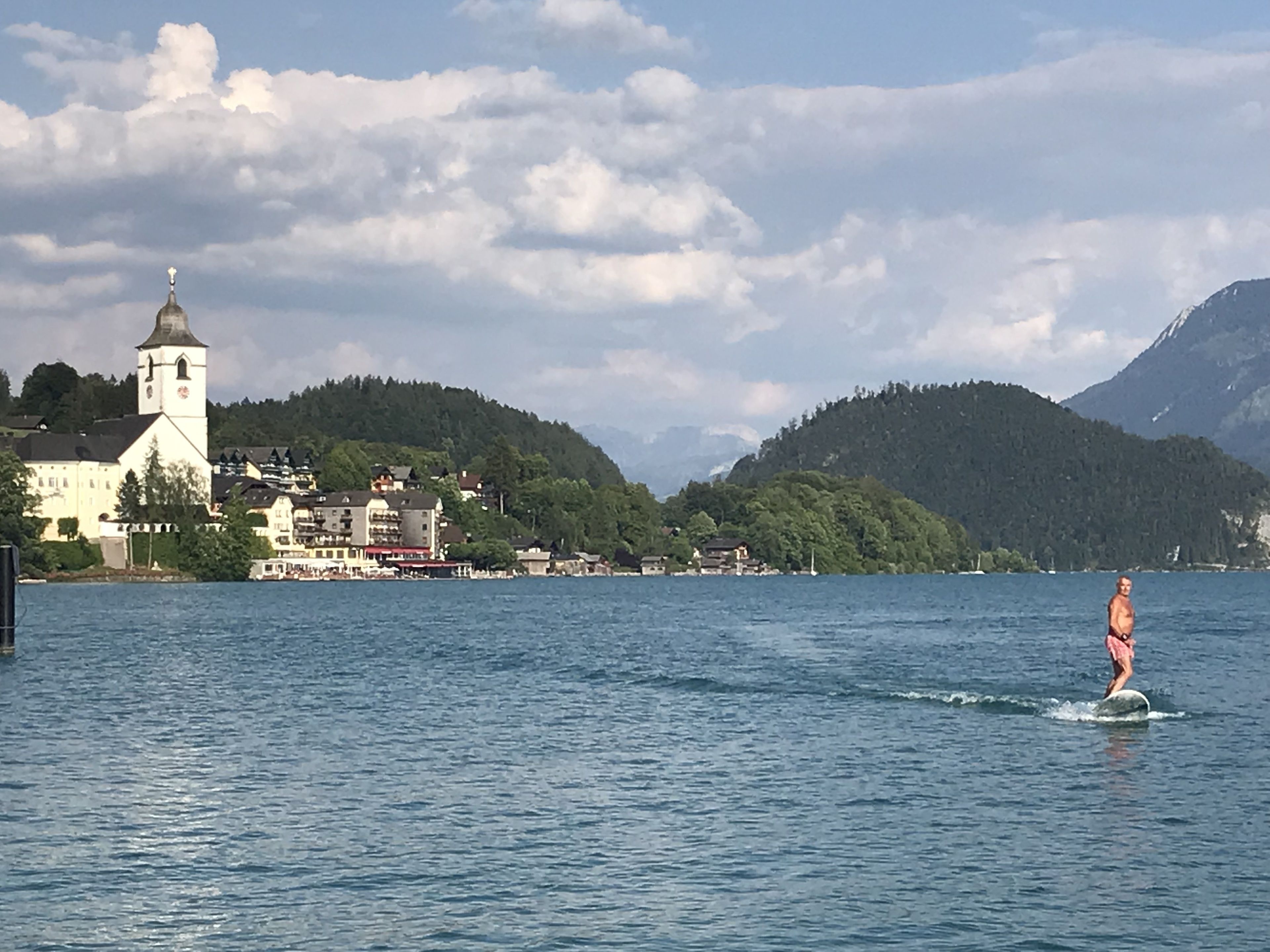 A lake with a church and buildings on the shore, a person wakeboarding, and mountains in the background.