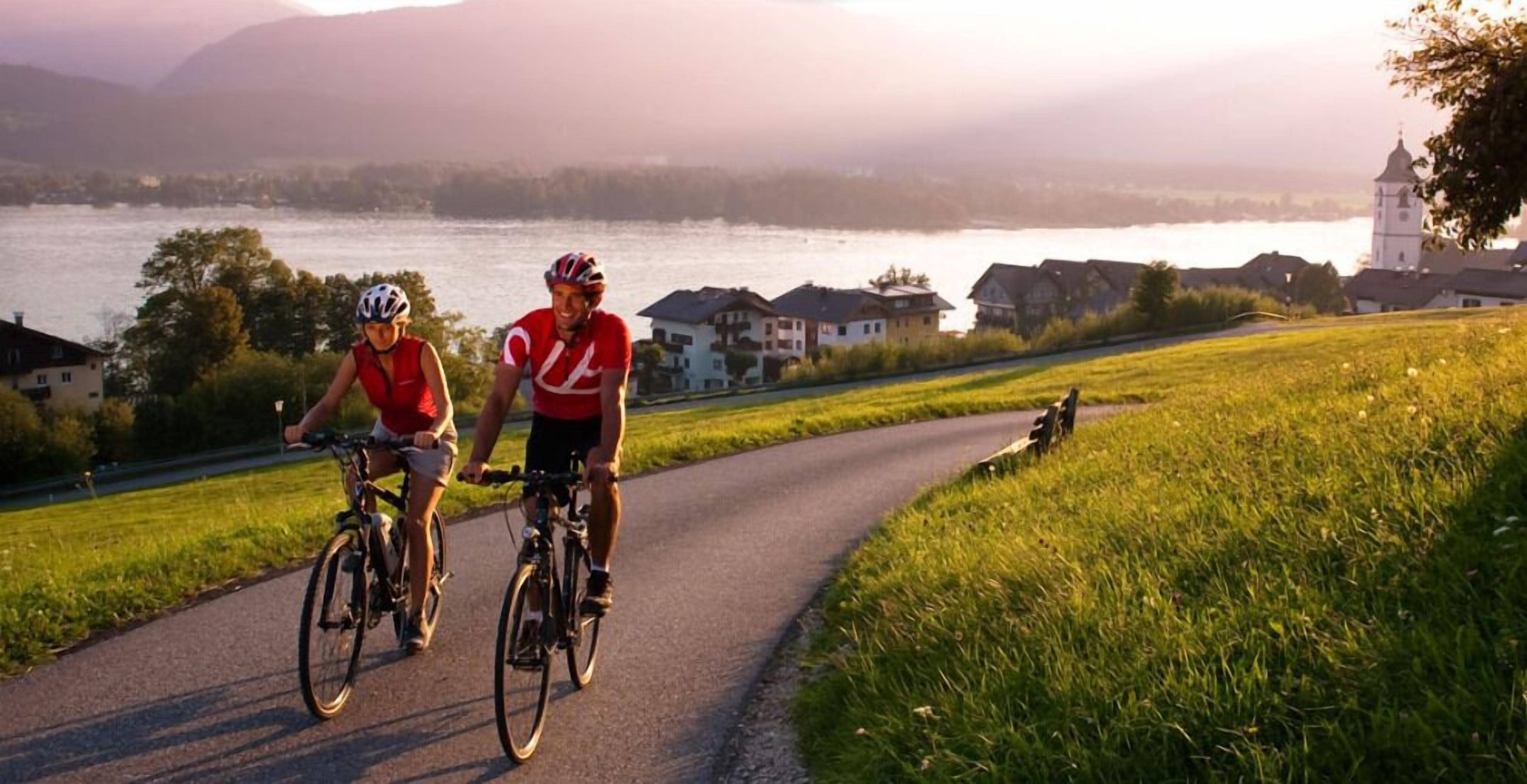 Two cyclists on a paved path with a lake and houses in the background.