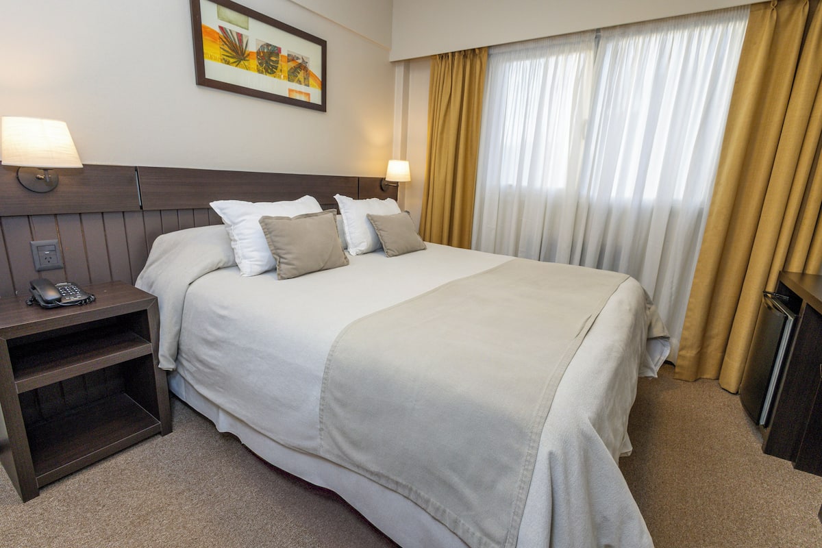 Une chambre d’hôtel avec un grand lit, deux lampes de chevet, une table de chevet avec un téléphone, un tableau encadré au mur et une fenêtre avec des rideaux.