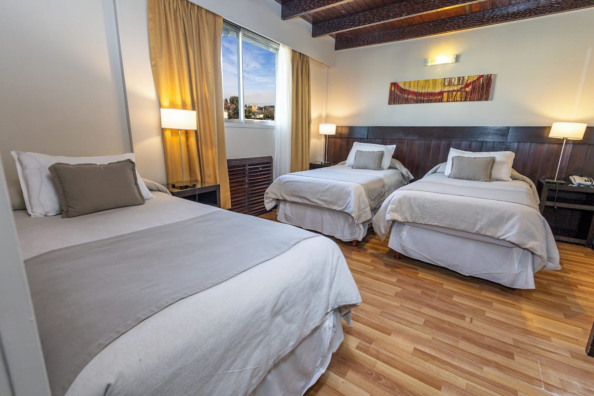 Une chambre d’hôtel avec deux lits simples, un plancher en bois, une fenêtre avec des rideaux, une table de chevet avec une lampe et un tableau au mur.