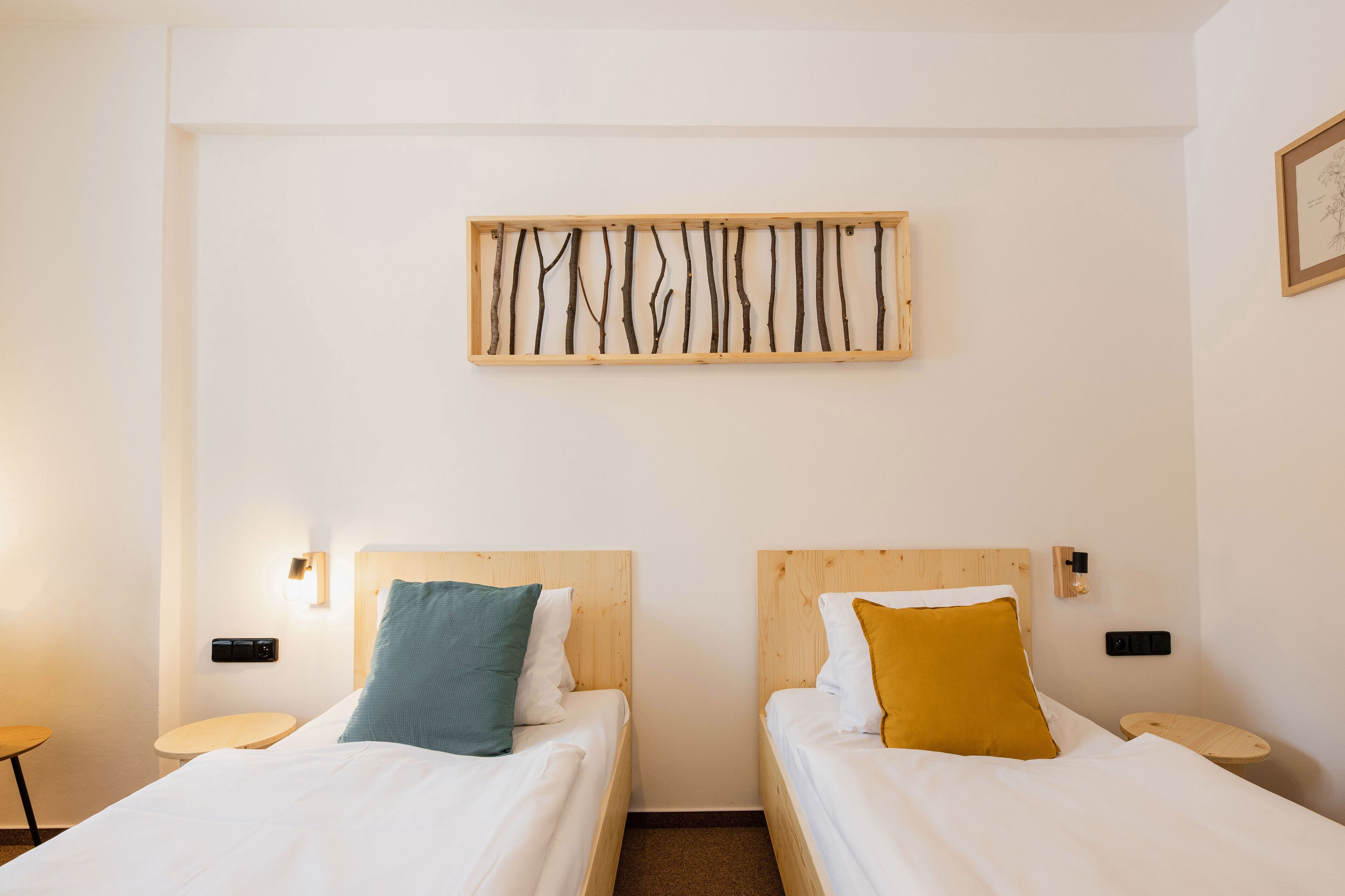 A hotel room with two single beds, each with a wooden headboard, white bedding, and a small bedside table. A wall-mounted light fixture and a framed picture are visible.