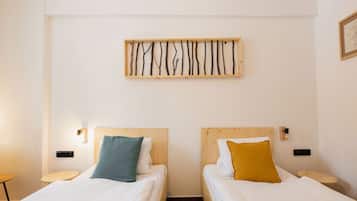 A hotel room with two single beds, each with a wooden headboard, white bedding, and a small bedside table. A wall-mounted light fixture and a framed picture are visible.