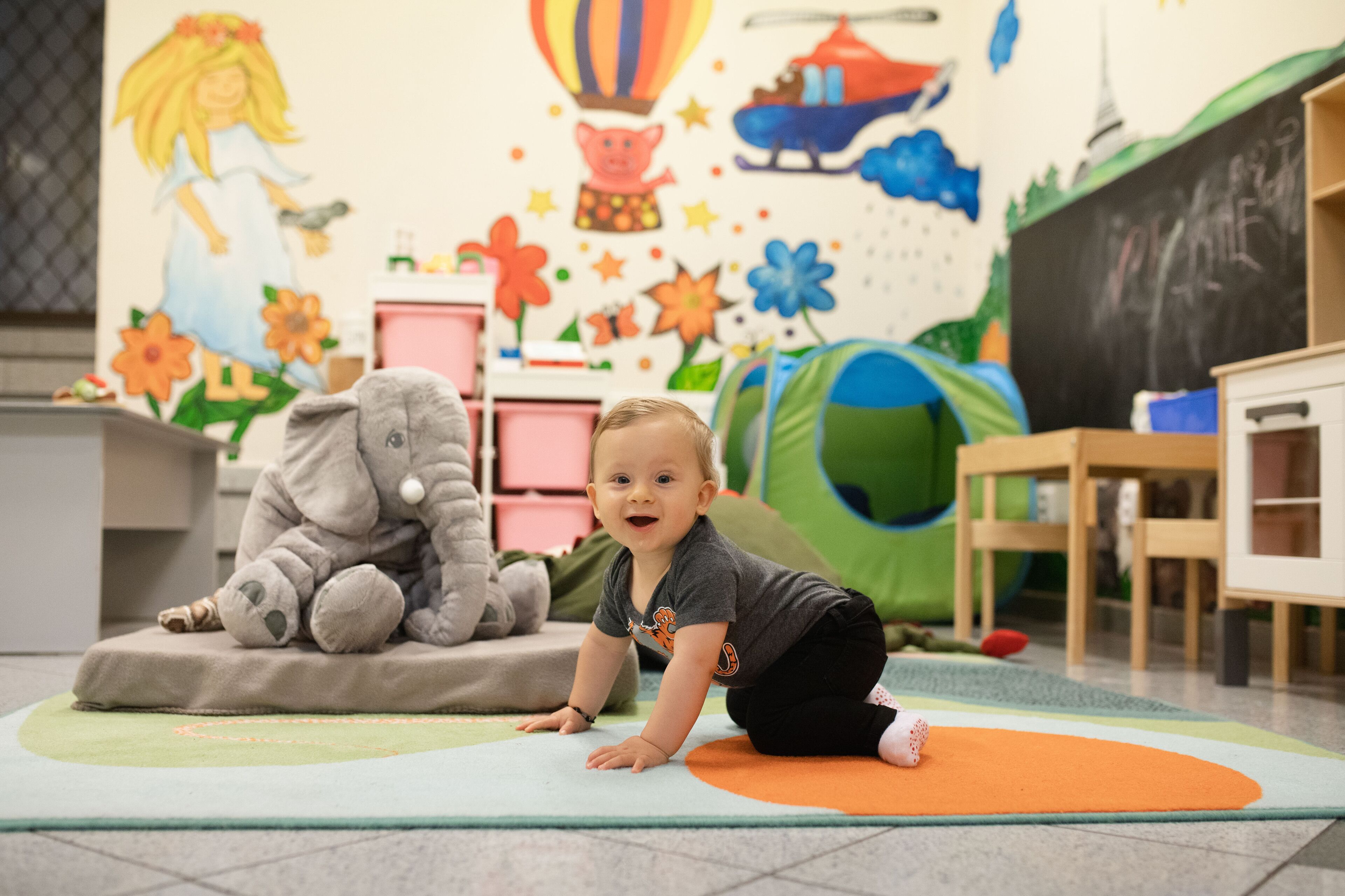 Ein Kind krabbelt auf einer bunten Spielmatte in einem Raum, dessen Wand mit Kinderbildern und einer Tafel verziert ist.