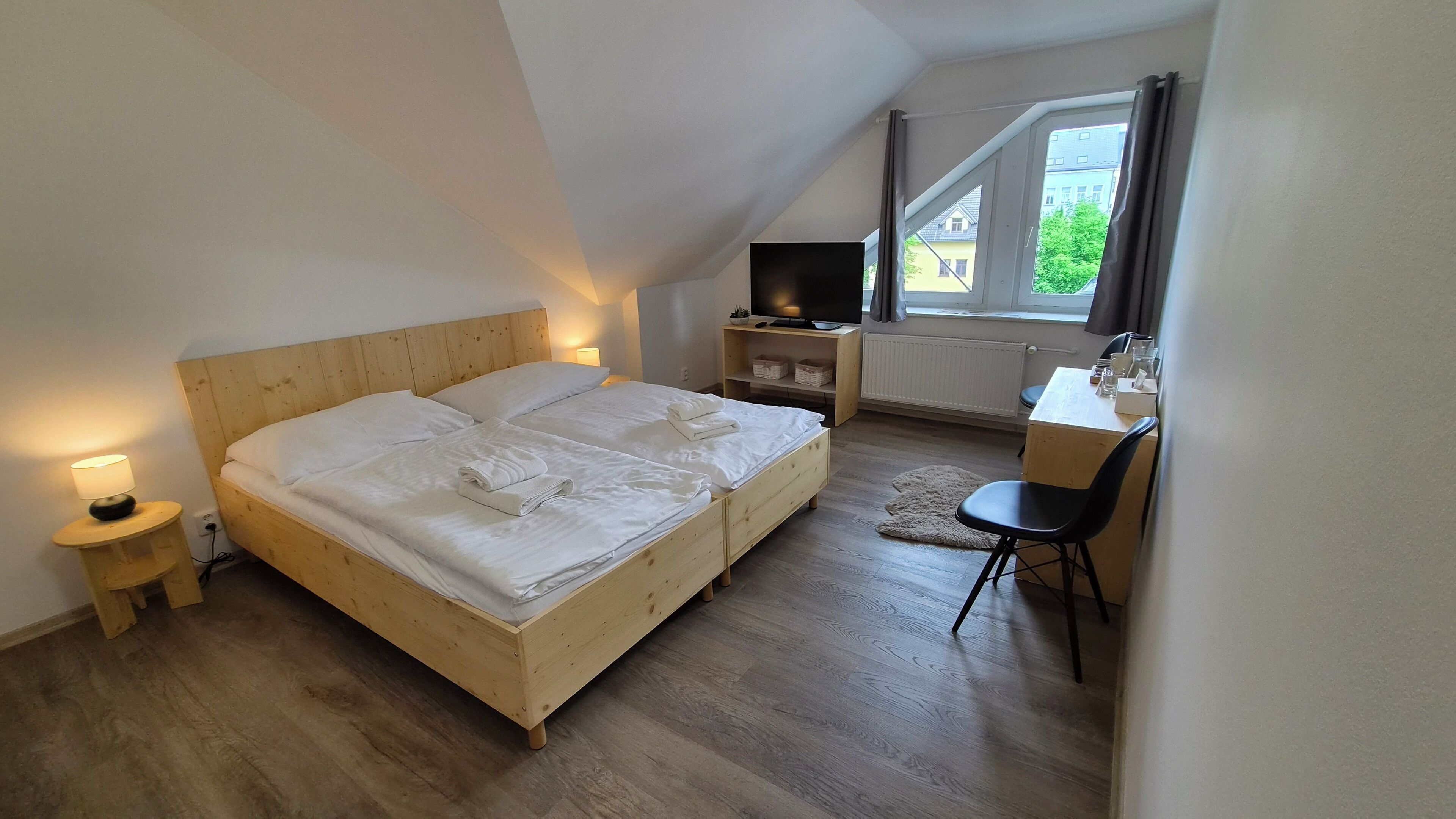 A bedroom with a wooden bed, bedside tables with lamps, a TV, a desk with a chair, and a view of buildings through a window.