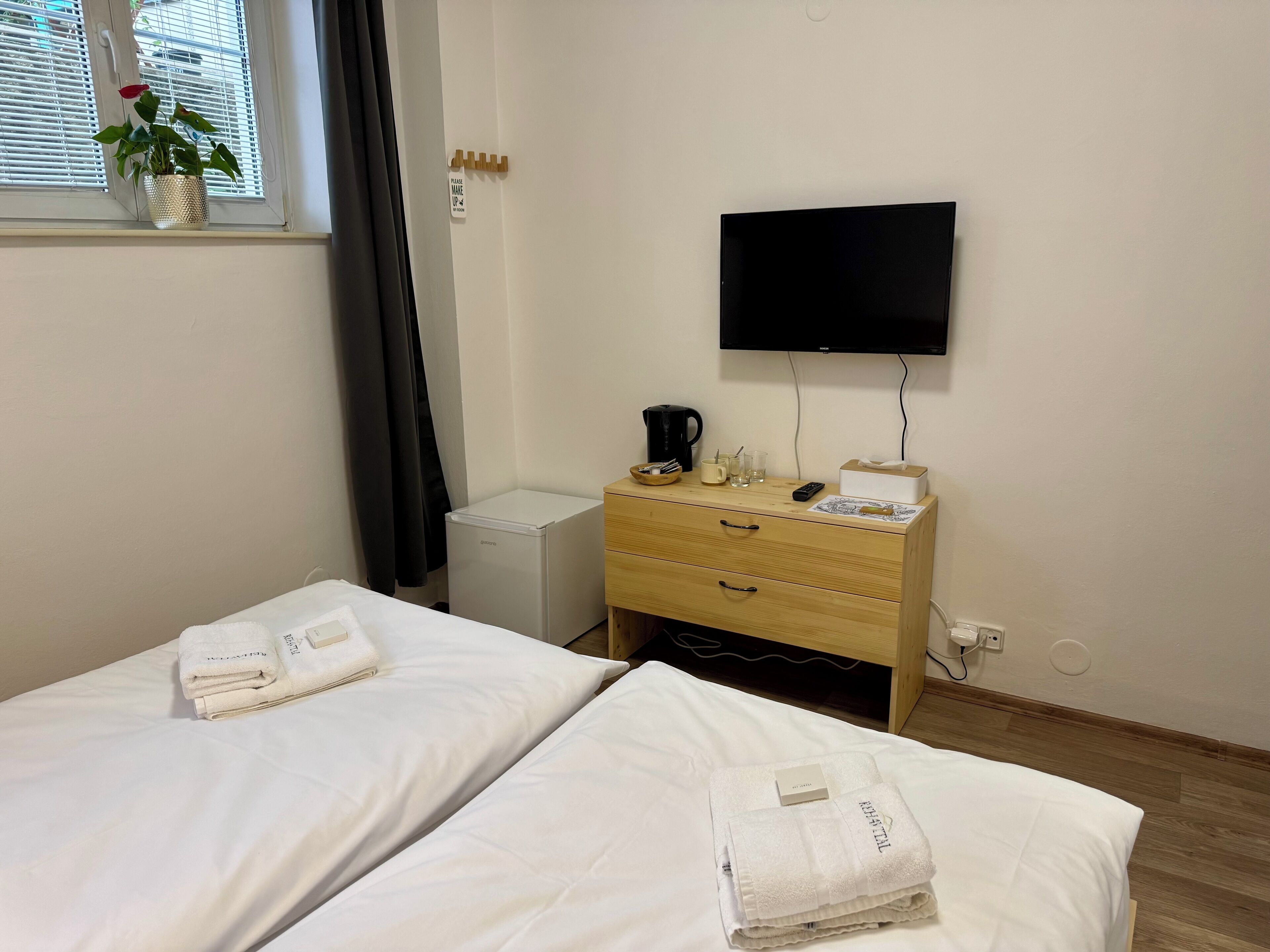 A hotel room with two beds, a TV, a small refrigerator, a wooden dresser, and a window with a plant.