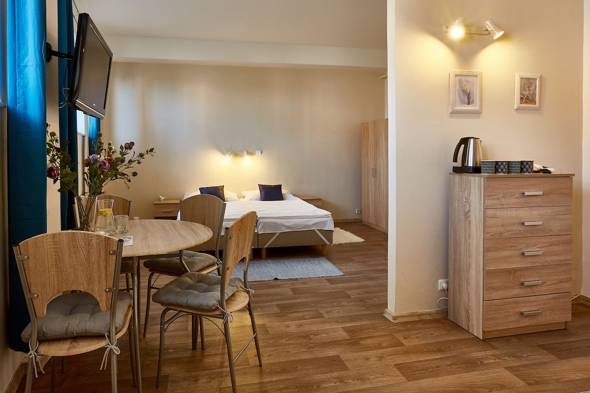 A compact room with a bed, a small table with chairs, a wooden cabinet, and a TV mounted on the wall.
