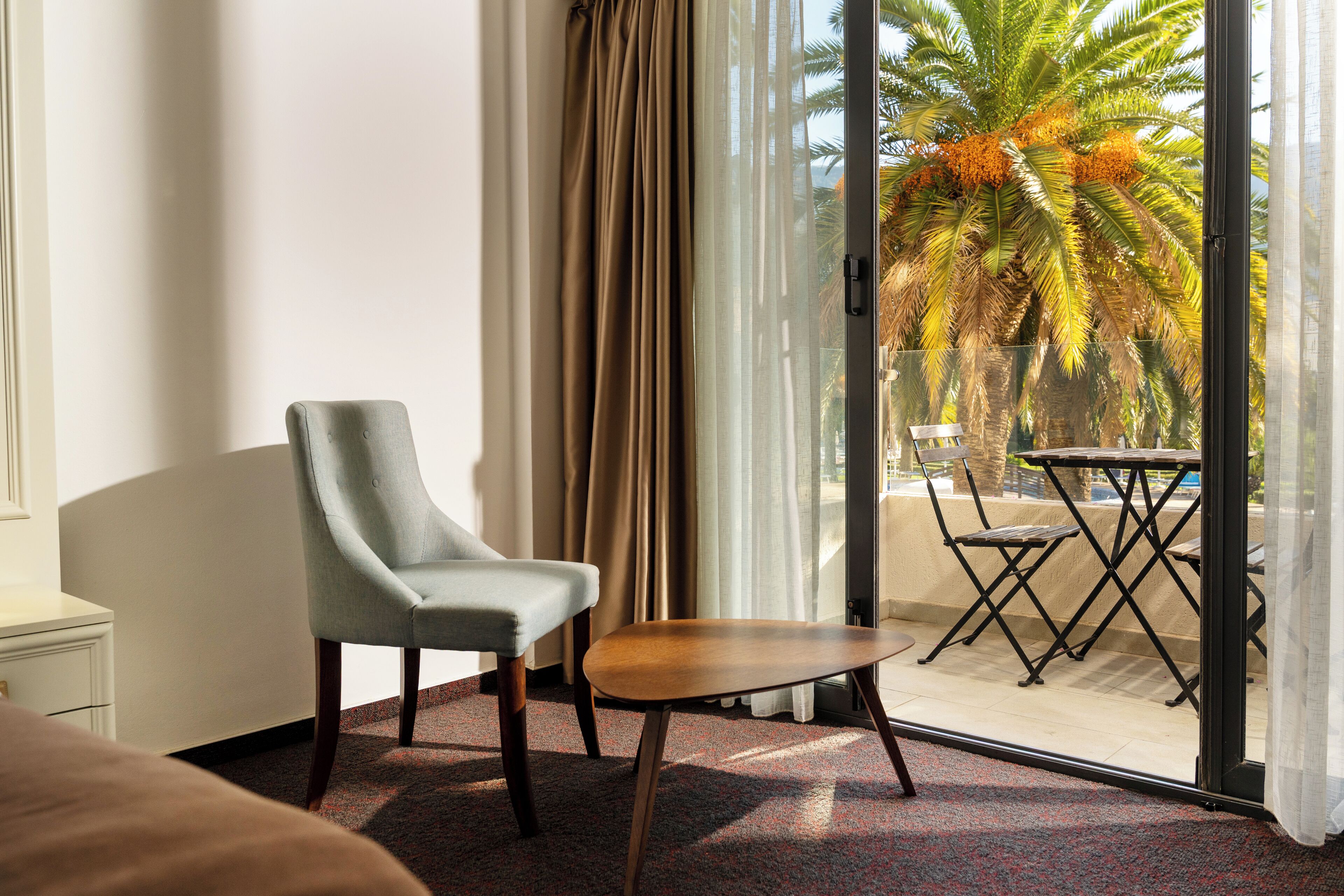 Habitación de hotel con una sola silla, una mesita y vistas a una palmera y a una zona de estar exterior a través de una puerta de cristal.