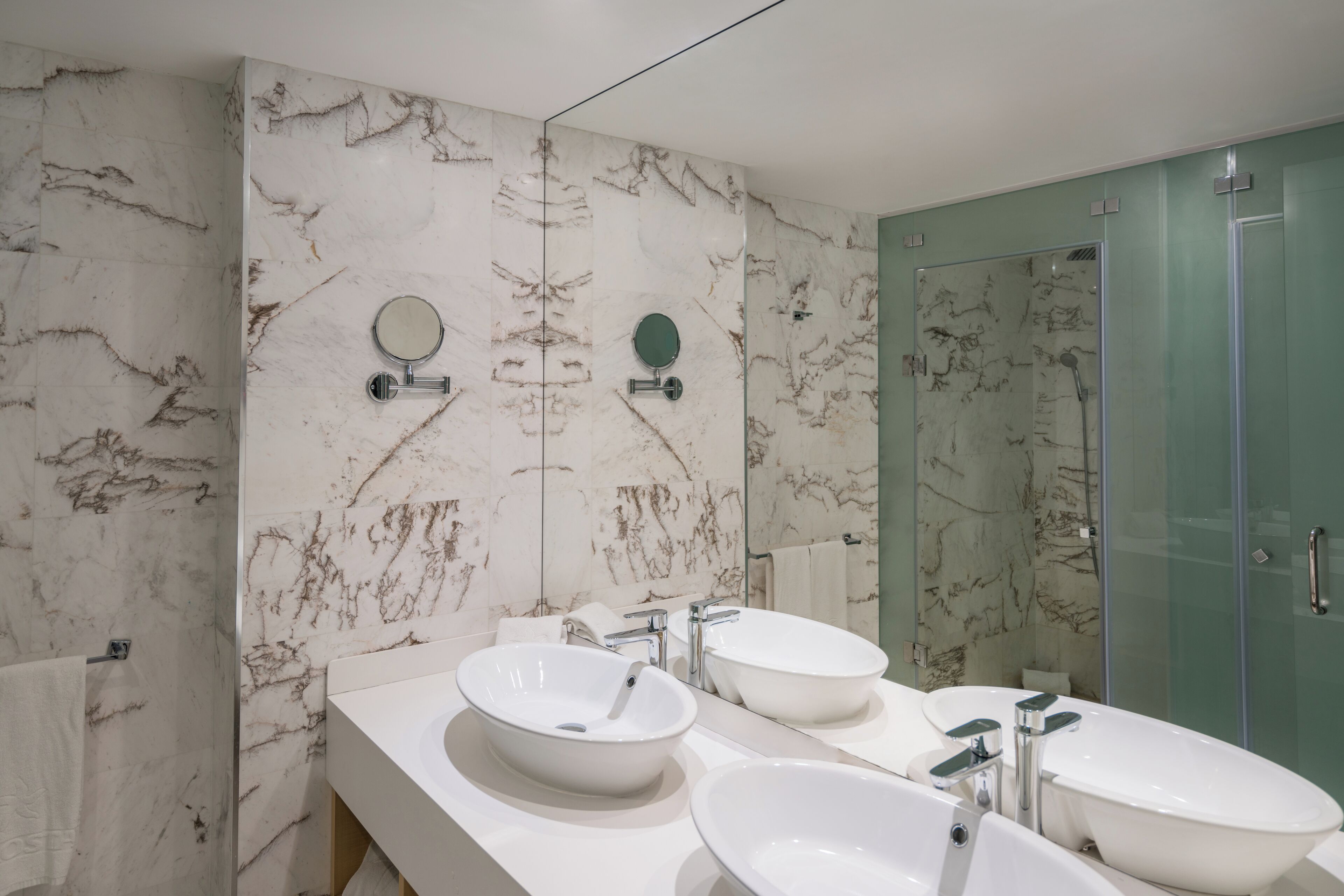 Une salle de bain moderne avec des murs en marbre, deux lavabos, un grand miroir et une douche à l’italienne en verre.