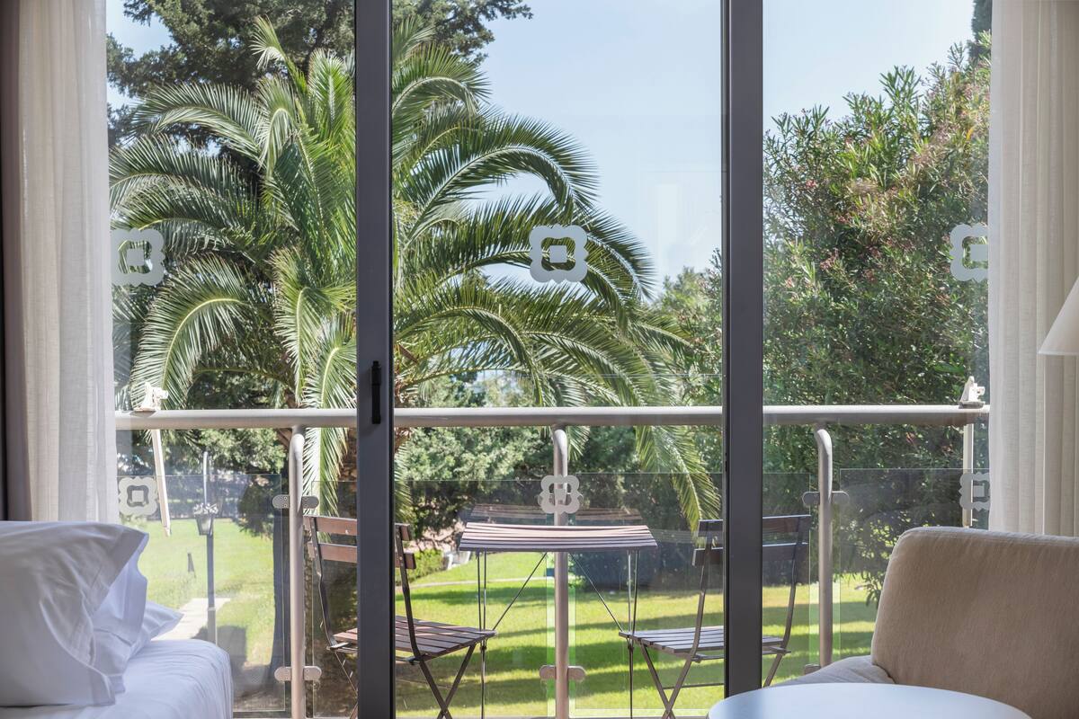A balcony with a glass railing, a bed, and a view of a garden with palm trees.