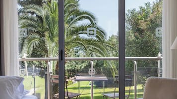 A balcony with a glass railing, a bed, and a view of a garden with palm trees.