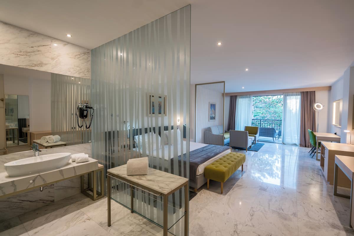 A modern hotel room with a large bathroom, a bed with a yellow ottoman, a desk, and a balcony with a view.