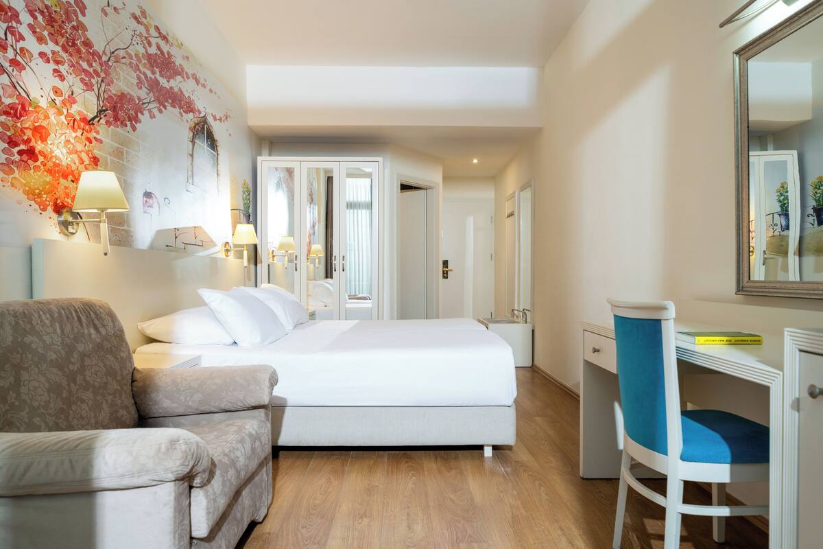 A bedroom with a bed, a desk, a chair, a sofa, and a wall with a floral mural.