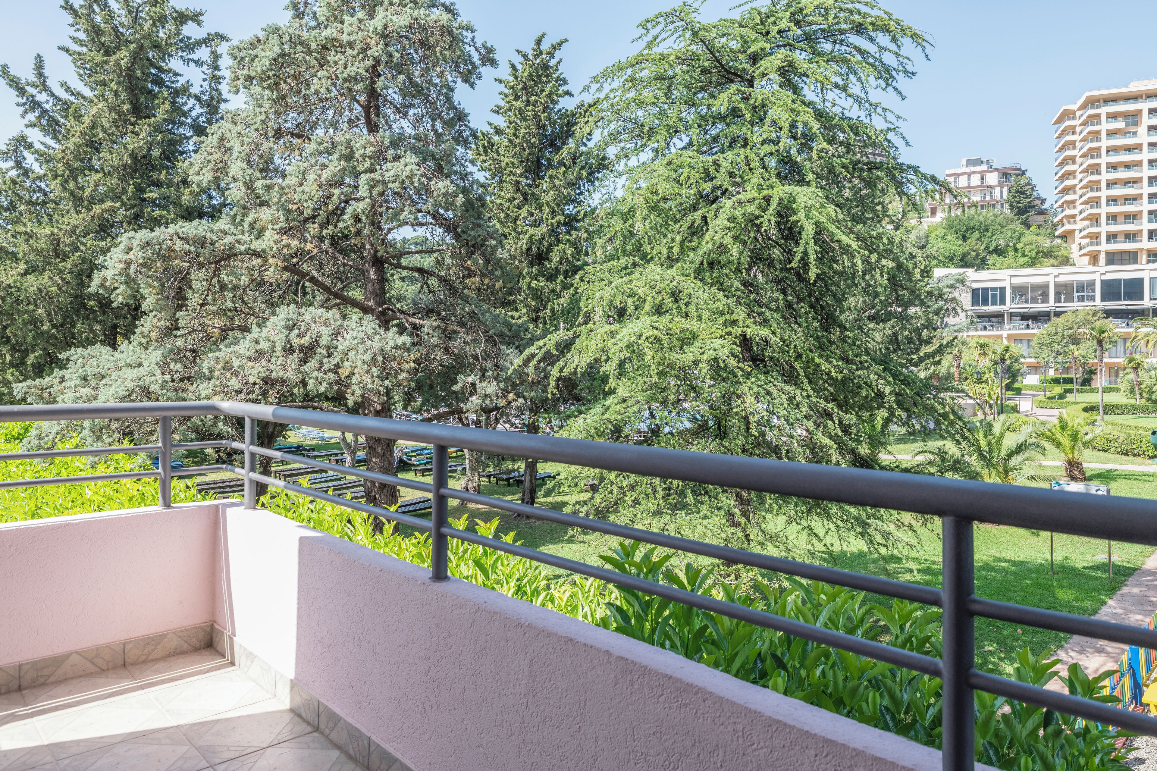 A balcony with a metal railing overlooking a garden with various trees and a building in the background.