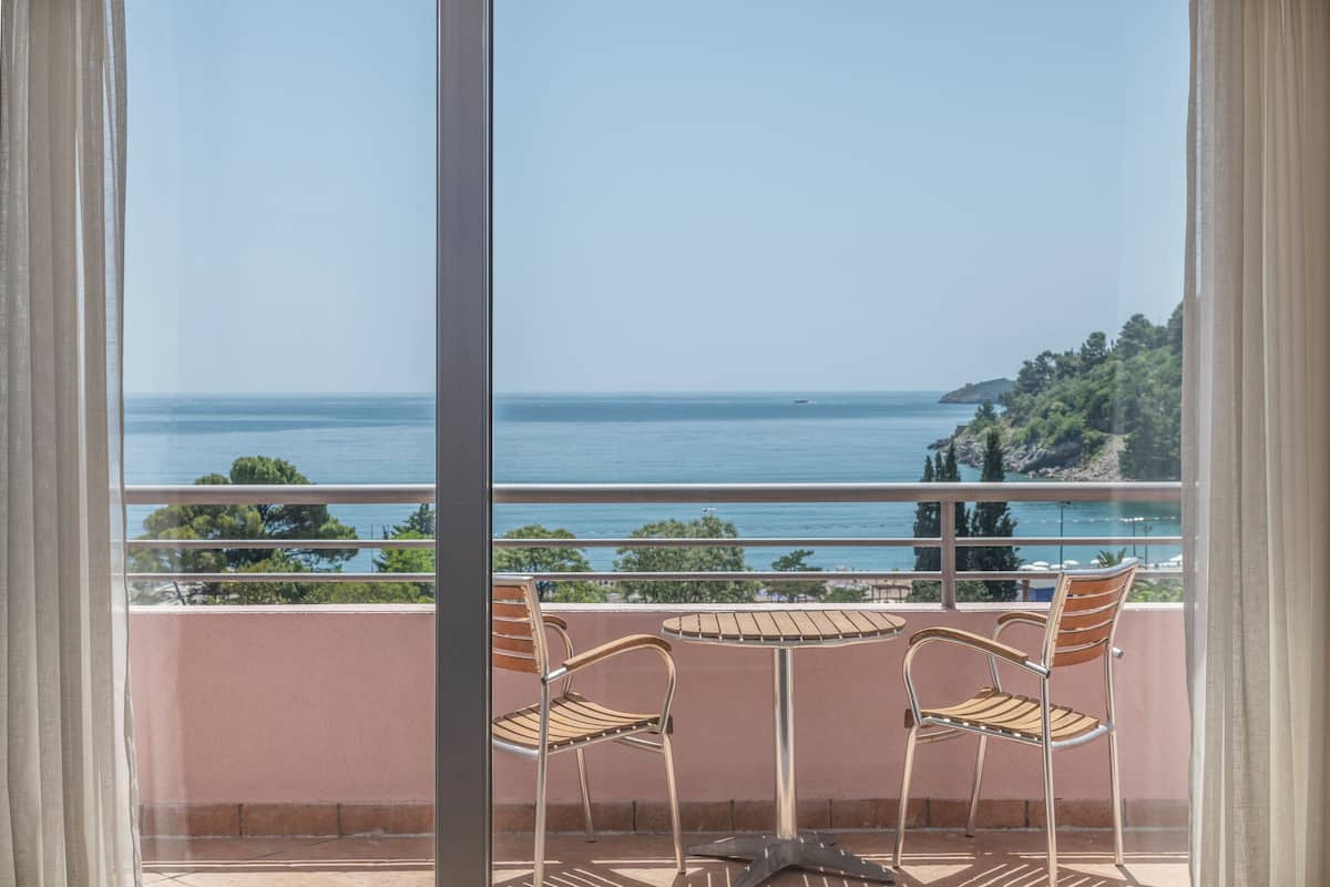 A balcony with two wooden chairs and a small table, overlooking a coastal view with trees and a clear sky.