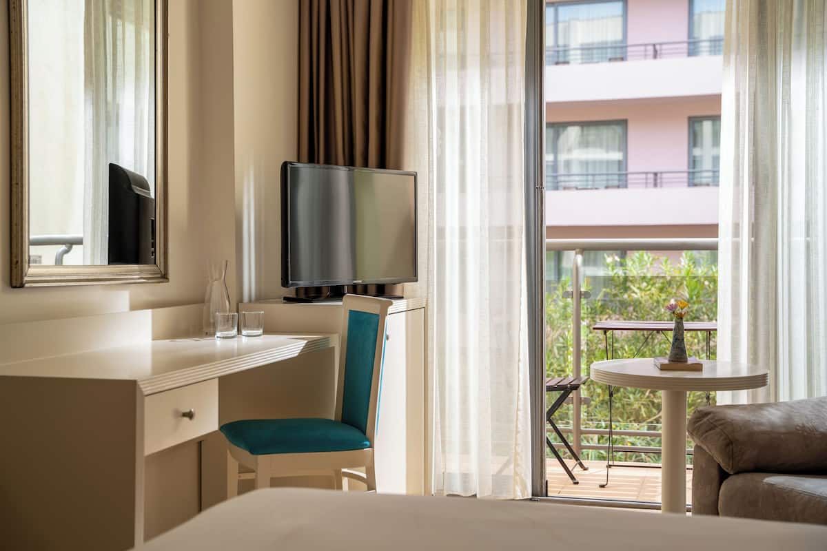 A hotel room with a bed, a desk with a chair, a television, a small table with a vase, and a balcony with a table and chairs.