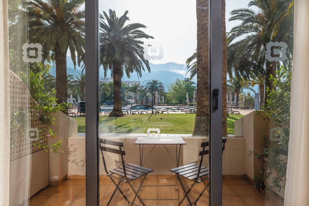 A balcony with two chairs and a table, overlooking a park with palm trees and a distant mountain.