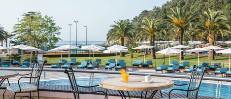 Zona junto a la piscina con sillas de descanso y sombrillas, una piscina y vistas al mar y al follaje.