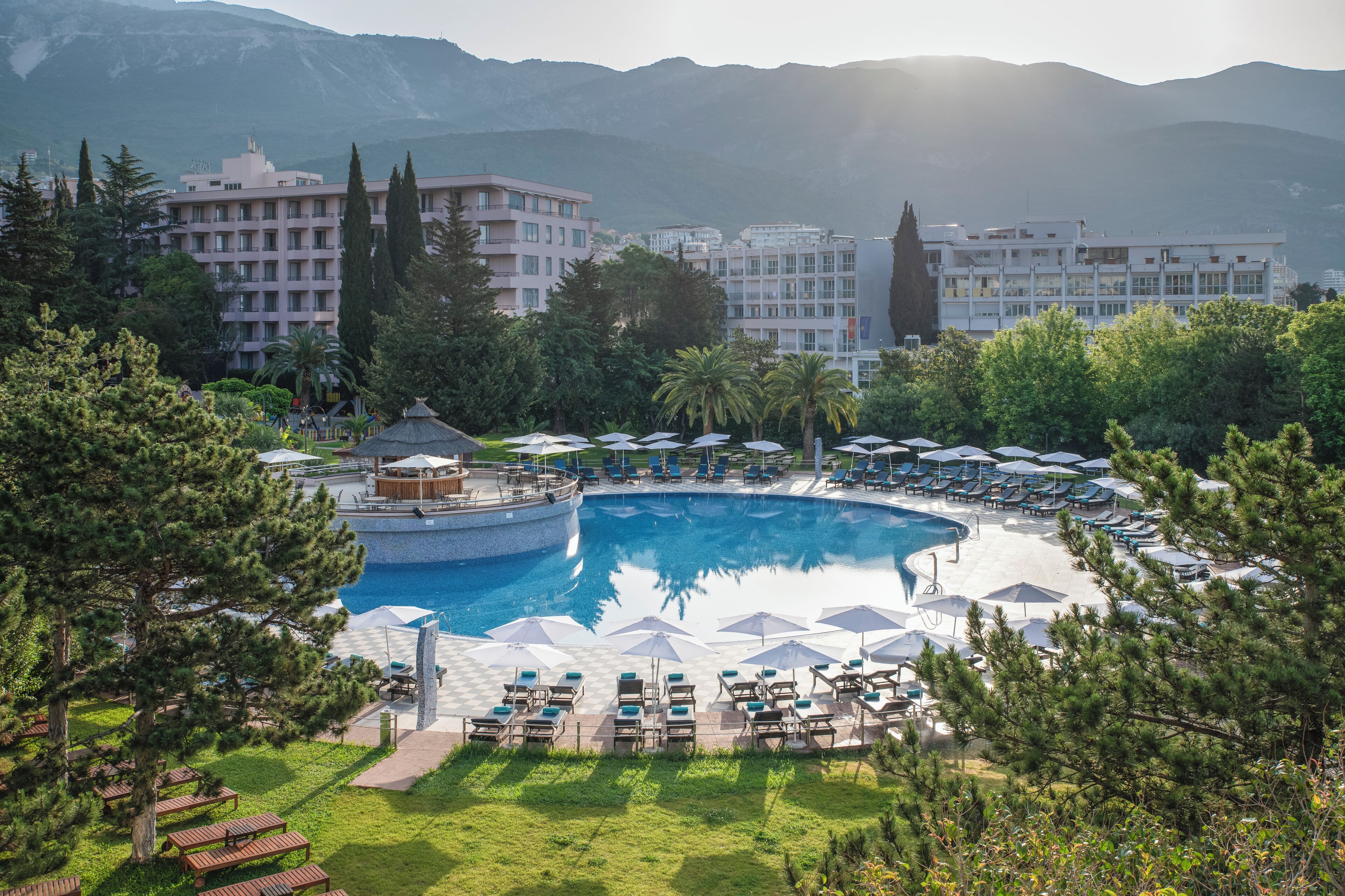 Un resort con una amplia piscina central rodeada de sillones de descanso y sombrillas, un pabellón y montañas al fondo.