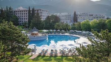Un resort con una amplia piscina central rodeada de sillones de descanso y sombrillas, un pabellón y montañas al fondo.
