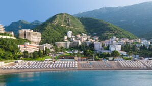 A coastal city with a beach area featuring rows of sun loungers and umbrellas, surrounded by buildings and mountains in the background.