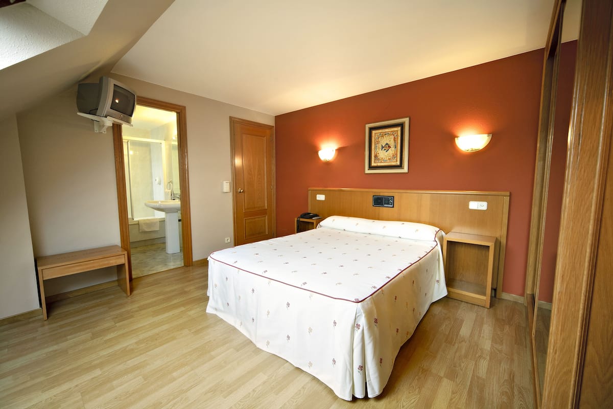Un dormitorio con cama, mesitas de noche, un banco, un televisor montado en la pared y un baño visible a través de una puerta abierta.