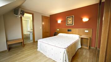 A bedroom with a bed, bedside tables, a bench, a television mounted on the wall, and a bathroom visible through an open door.