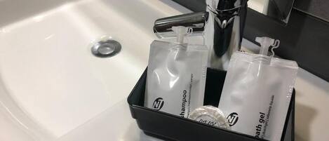Uma pia de banheiro com torneira cromada e um saboneteira preta com duas garrafas brancas rotuladas como “shampoo” e “gel de banho”.