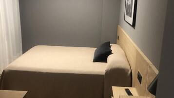 A small, neatly arranged room with a single bed, a bedside table, a chair, and a desk.