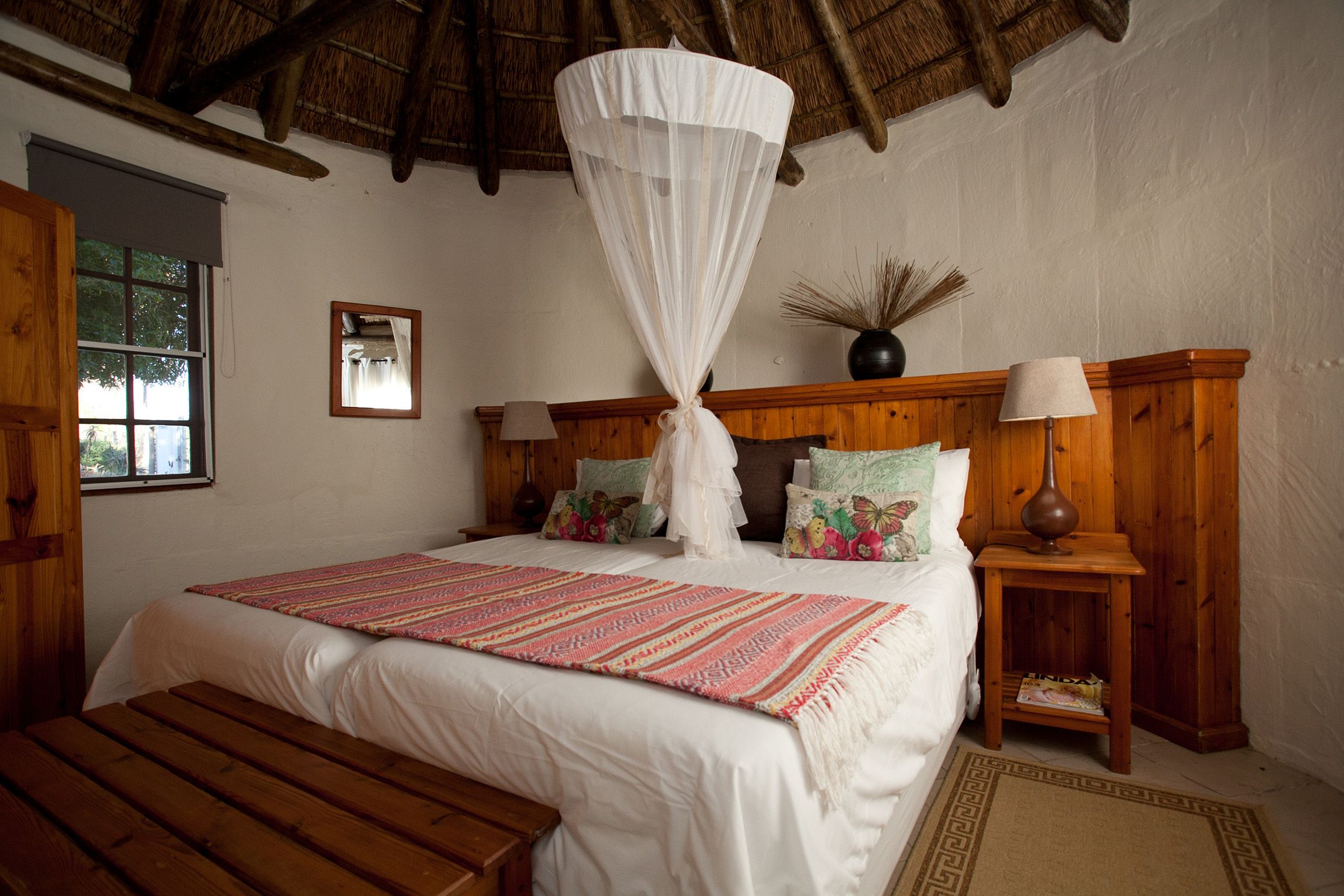A bedroom with a large bed, wooden headboard, bedside tables with lamps, a window, and a thatched roof.