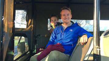 A vehicle interior with two people seated, one in a blue jacket and the other in a brown jacket, with a window showing greenery outside.