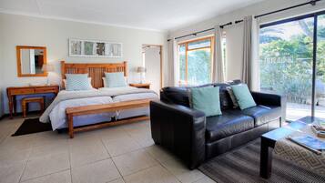 Un dormitorio con cama, un sofá de cuero negro, un banco de madera, una mesita de noche, una lámpara y un amplio ventanal con cortinas.