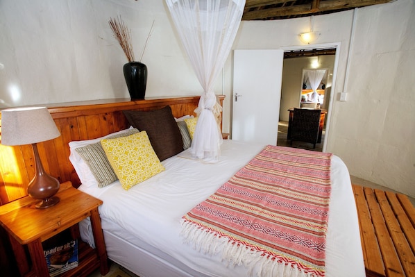 A neatly made bed with a white comforter, a patterned throw blanket, and a wooden headboard. There is a bedside table with a lamp and a vase with dried plants.