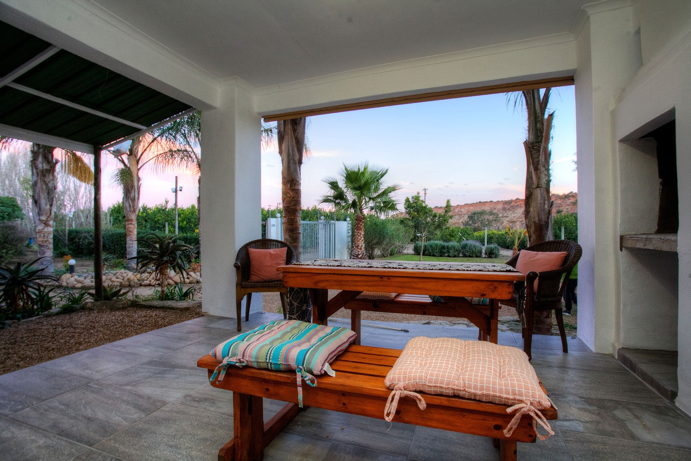 An outdoor patio with a wooden table and benches, surrounded by palm trees and a view of a desert landscape.