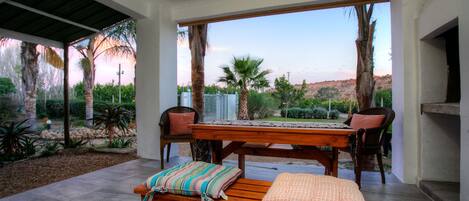 An outdoor patio with a wooden table and benches, surrounded by palm trees and a view of a desert landscape.