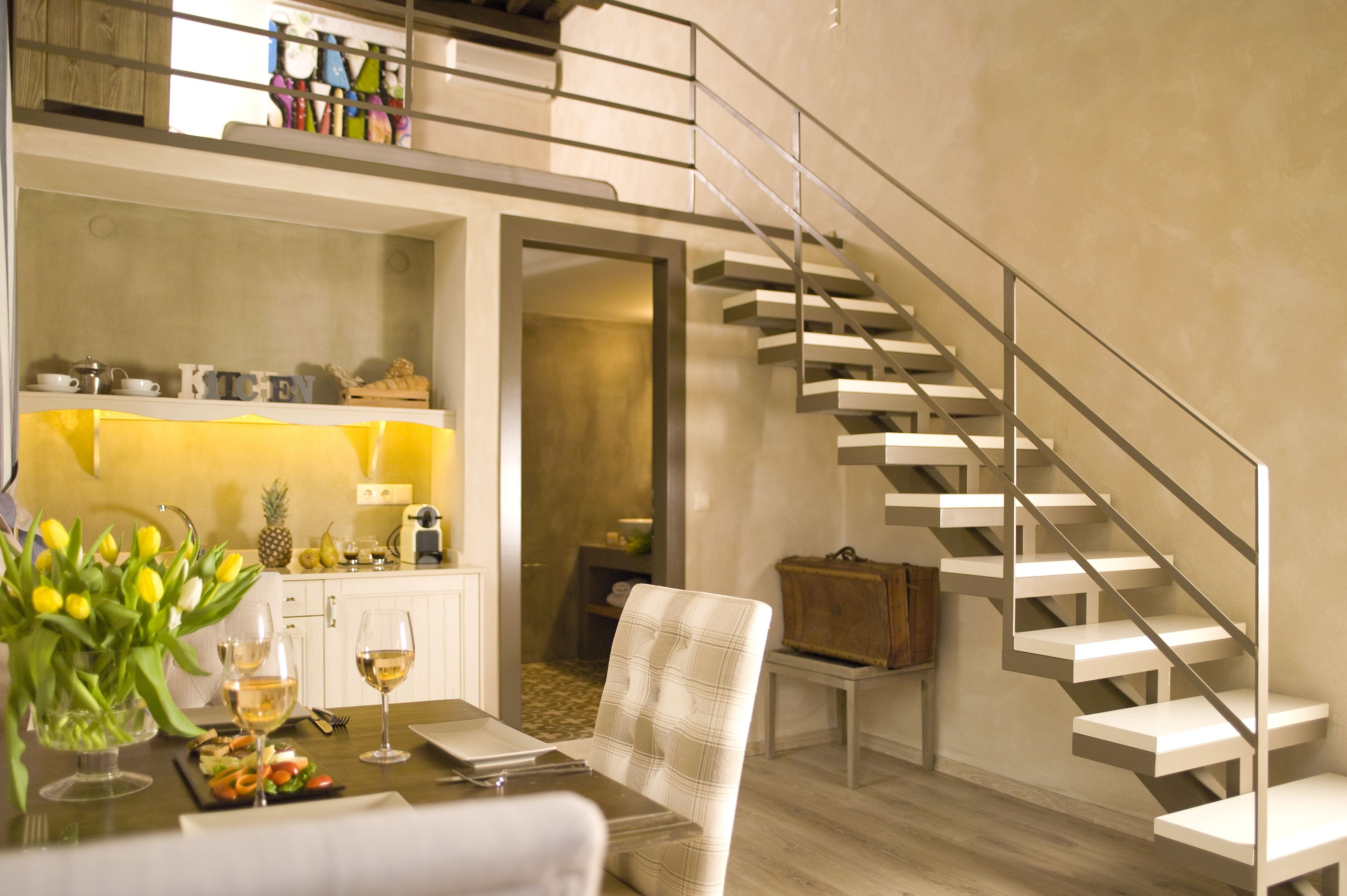 Una cocina moderna con armarios blancos, un comedor con una mesa dispuesta para dos personas, y una escalera de diseño minimalista.