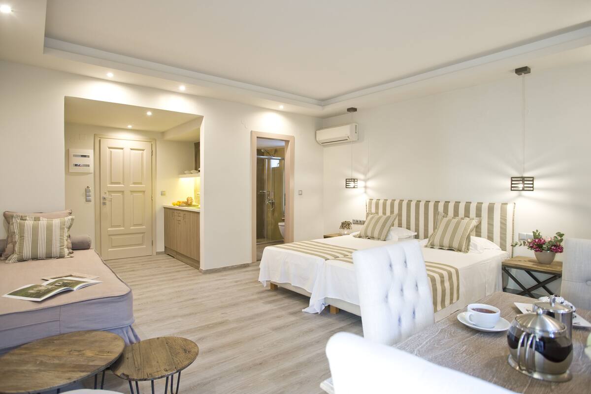 A modern hotel room with two beds, a sitting area with a table and chairs, and a kitchenette.
