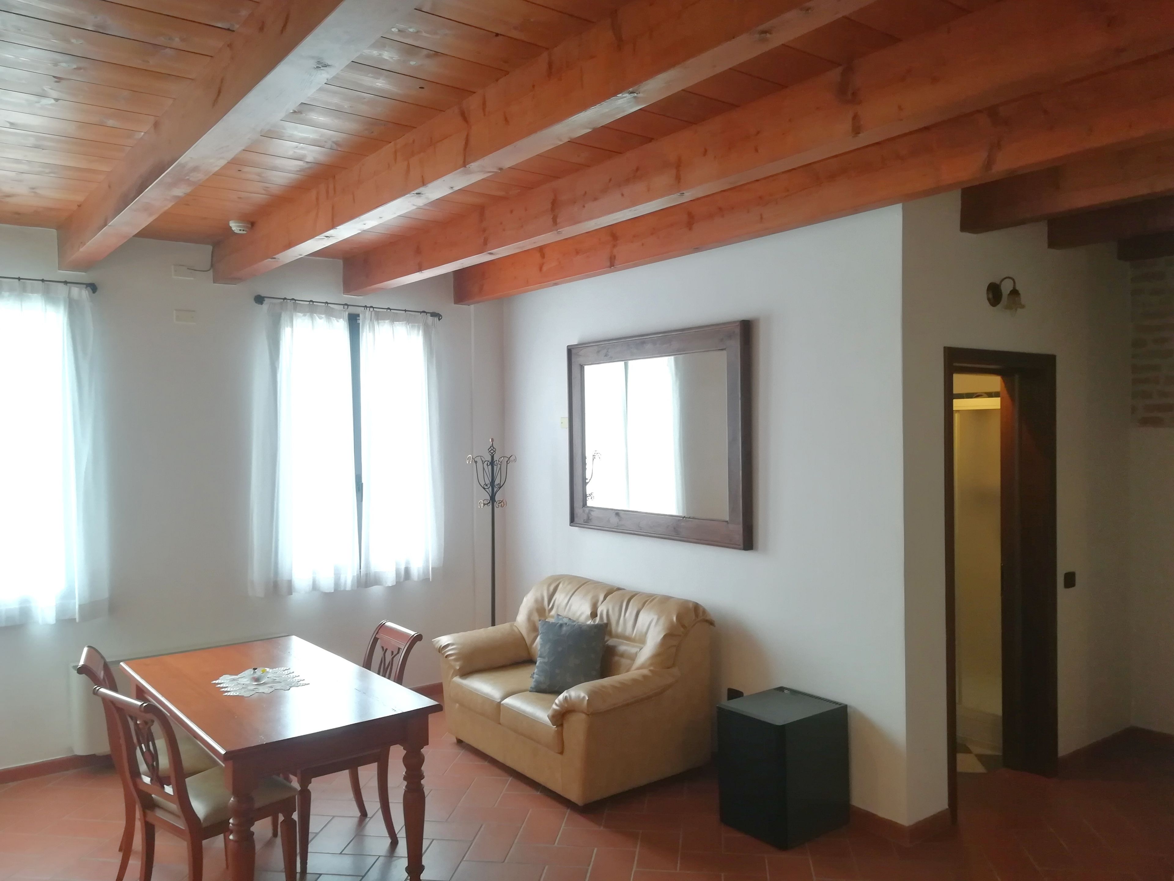 A room with a wooden ceiling, a dining table with chairs, a sofa, a small black speaker, and a mirror on the wall.